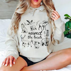 Puede incluir: Una sudadera con capucha de color crema con un gr&aacute;fico negro de varios animales del bosque, incluyendo un lobo, un mapache y un oso. La frase "respect the locals" est&aacute; escrita en un estilo manuscrito debajo de las ilustraciones de animales. La sudadera tiene una capucha con cord&oacute;n y un bolsillo de canguro.