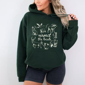 Puede incluir: Sudadera con capucha verde oscuro con bolsillo de canguro. La parte delantera presenta un gr&aacute;fico blanco de varios animales y el texto "respect the locals". El dise&ntilde;o incluye un lobo, un ciervo, un oso, un conejo y otros animales salvajes.