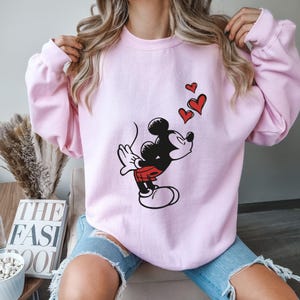 K&ouml;nnte beinhalten: Hellrosa Sweatshirt mit einer Grafik von Mickey Mouse, der drei rote Herzen pustet. Mickey ist in seiner klassischen schwarzen, roten und wei&szlig;en Kleidung dargestellt. Das Sweatshirt hat lange &Auml;rmel und einen Rundhalsausschnitt.