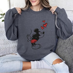 K&ouml;nnte beinhalten: Dunkelgraues Sweatshirt mit einer Grafik von Micky Maus, die K&uuml;sse pustet, mit roten Herzen, die dar&uuml;ber schweben. Micky ist in Schwarz, mit roten Shorts und wei&szlig;en Handschuhen dargestellt. Das Sweatshirt hat lange &Auml;rmel und einen Rundhalsausschnitt.