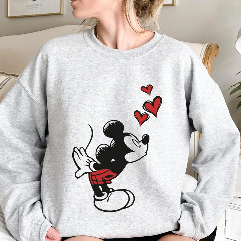 K&ouml;nnte beinhalten: Ein hellgraues Sweatshirt mit einer Grafik von Micky Maus, die K&uuml;sse pustet, mit drei roten Herzen, die dar&uuml;ber schweben. Micky ist in seiner klassischen schwarzen, roten und wei&szlig;en Kleidung dargestellt. Das Sweatshirt hat lange &Auml;rmel und einen Rundhalsausschnitt.