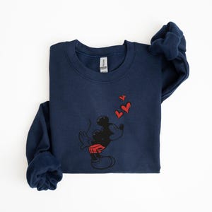 K&ouml;nnte beinhalten: Marineblaues Sweatshirt mit einem schwarzen Cartoon-Charakter, der rote herzf&ouml;rmige K&uuml;sse pustet. Die Figur tr&auml;gt rote Shorts. Das Sweatshirt hat einen Rundhalsausschnitt und lange &Auml;rmel.