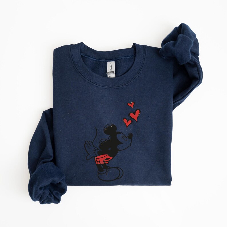 K&ouml;nnte beinhalten: Marineblaues Sweatshirt mit einem schwarzen Cartoon-Charakter, der rote herzf&ouml;rmige K&uuml;sse pustet. Die Figur tr&auml;gt rote Shorts. Das Sweatshirt hat einen Rundhalsausschnitt und lange &Auml;rmel.