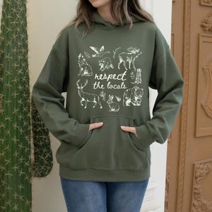 Puede incluir: Una sudadera con capucha verde oliva con bolsillo de canguro y capucha. La parte delantera de la sudadera con capucha presenta un gr&aacute;fico blanco de varios animales y el texto "respect the locals". Los animales incluyen un lobo, un oso, un ciervo, un conejo y otros animales salvajes.