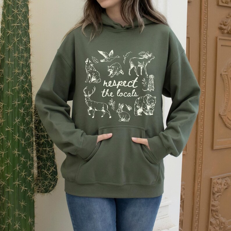 Puede incluir: Una sudadera con capucha verde oliva con bolsillo de canguro y capucha. La parte delantera de la sudadera con capucha presenta un gr&aacute;fico blanco de varios animales y el texto "respect the locals". Los animales incluyen un lobo, un oso, un ciervo, un conejo y otros animales salvajes.