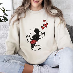 K&ouml;nnte beinhalten: Cremefarbenes Sweatshirt mit einer Grafik von Micky Maus, die K&uuml;sse pustet, mit drei roten Herzen dar&uuml;ber. Micky ist in seinem klassischen Outfit dargestellt: schwarzer K&ouml;rper, rote Shorts, wei&szlig;e Handschuhe und Schuhe. Das Sweatshirt ist ein l&auml;ssiger, bequemer Stil.