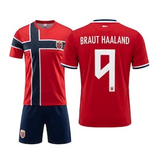 May include: A red soccer jersey and shorts set featuring a dark blue and white cross pattern on the front, the Norway flag emblem, and the back displaying the name BRAUT HAALAND with the number 9 in white lettering on the red fabric.