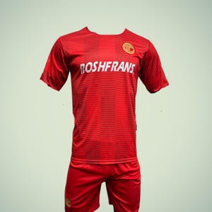 May include: Red soccer uniform with the text "ROSHFRANS" across the chest. The jersey has a striped pattern and a circular emblem on the upper right. Matching red shorts complete the set. The uniform is designed for sports.