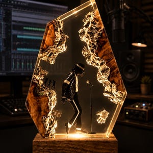 May include: A decorative resin and wood lamp featuring a miniature figure of a performer in a black suit and hat, standing next to a microphone. The piece includes a small glove icon, a sign, and silhouettes, all encased in a clear, angular resin block.