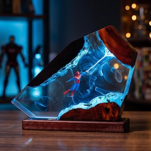 May include: A decorative lamp featuring a miniature Spider-Man figure suspended within a clear resin block. The resin is framed by natural wood edges and illuminated with a bright blue light, creating a swirling smoke-like effect inside.