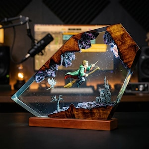 Puede incluir: Escultura decorativa de resina que representa a un niño en una escoba, un castillo y un pájaro blanco. La escultura está encerrada en resina transparente con una base y detalles de madera. El niño lleva una capa verde y sostiene una varita. La escultura mide unos 20 cm de altura.