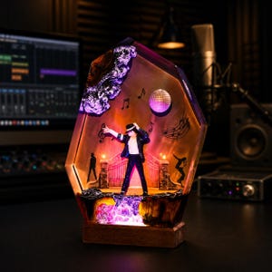 May include: A hexagonal resin display piece featuring a miniature figure of a performer in a black suit and white shirt, standing before a gate. The scene includes a disco ball, musical notes, and silhouettes, illuminated with orange and purple light.