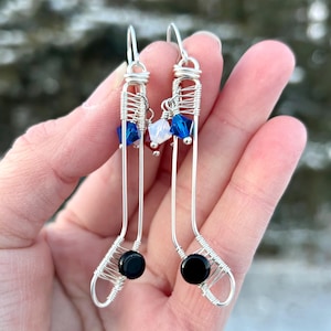 May include: A pair of silver wire earrings shaped like hockey sticks with black beads at the end of each stick. The earrings are decorated with red, white, and blue crystals.