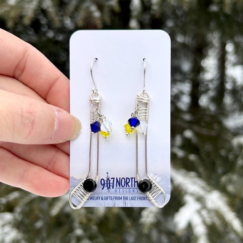 May include: Silver wire earrings with black, blue, yellow, and white crystal beads. The earrings are shaped like hockey sticks. The earrings are on a white card with the text "947 North Designs Jewelry & Gifts from the Last Frontier."