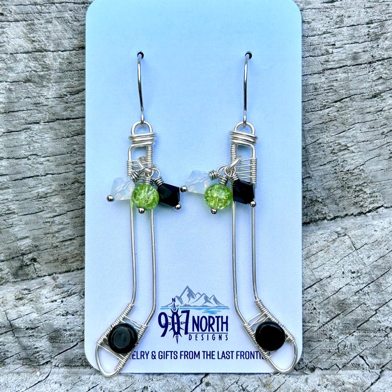 May include: Silver wire earrings with green and black beads, shaped like a sock. The earrings are on a white card with the text "907 North Designs Jewelry & Gifts from the Last Frontier".