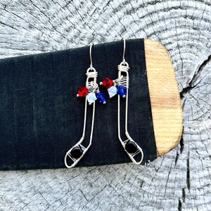 May include: A pair of silver wire earrings shaped like hockey sticks with red, white, and blue crystal beads. The earrings have black beads at the bottom.