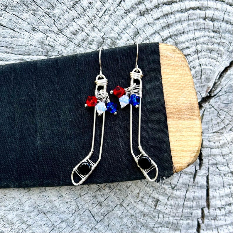 May include: A pair of silver wire earrings shaped like hockey sticks with red, white, and blue crystal beads. The earrings have black beads at the bottom.