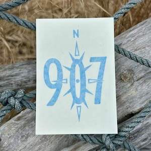 May include: A blue and white sticker with the number 907 and a compass rose design. The compass rose has a black dot in the center and points in all four directions.