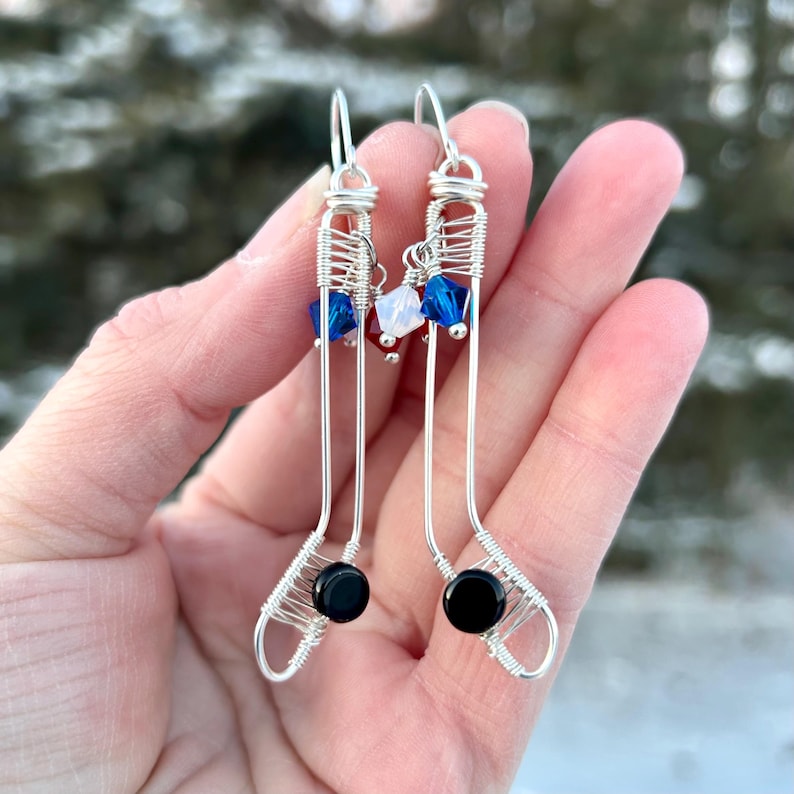 May include: A pair of silver wire earrings shaped like hockey sticks with black beads at the end of each stick. The earrings are decorated with red, white, and blue crystals.
