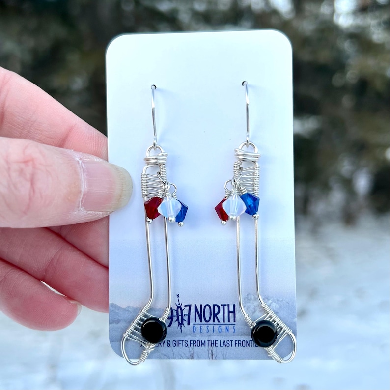 May include: Silver wire earrings shaped like hockey sticks with red, white, and blue crystal beads and black beads at the bottom. The earrings are on a white card with the text "#7 North Designs" and "Jewelry & Gifts from the Last Frontier".