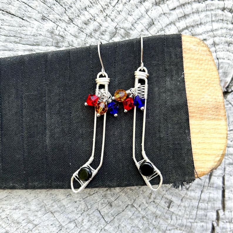 May include: A pair of silver wire earrings shaped like hockey sticks with black and clear beads at the bottom and red, orange, and blue crystal beads at the top.