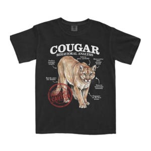 Könnte beinhalten: Ein schwarzes T-Shirt mit kurzen Ärmeln und einer Grafik eines Pumas. Der Text lautet COUGAR BEHAVIORAL ANALYSIS, mit Beschriftungen, die auf das Tier zeigen, und einem roten kreisförmigen Stempel mit der Aufschrift PROCEED WITH CAUTION.