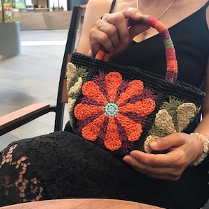 May include: A black crocheted handbag featuring a large central flower motif with orange and purple petals and a light blue center. The bag has a multi-colored striped handle and decorative beige leaf patterns on the sides, all crafted from yarn.