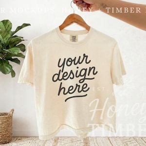 May include: Cream-colored t-shirt with the text "your design here" in a black, cursive font. The shirt is hanging on a wooden hanger. A potted plant is in the background.