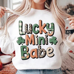 Puede incluir: Sudadera blanca con el texto "Lucky Mini Babe" en una mezcla de fuentes y patrones, incluyendo cuadros, estampado de leopardo y colores lisos. También se incluyen gráficos de tréboles verdes.