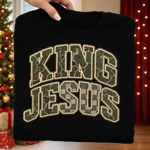 May include: Black sweatshirt with the words "KING JESUS" in a camouflage and checkered design. The text is outlined in a tan color. The sweatshirt is held up by a hand.