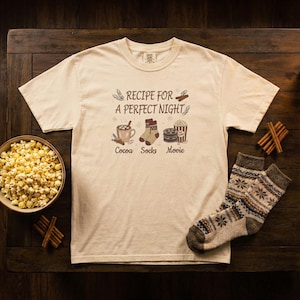 May include: Cream-colored t-shirt with the text "Recipe for a Perfect Night" and illustrations of cocoa, socks, and a movie. A bowl of popcorn and a pair of patterned socks are also visible. Cinnamon sticks are placed on the wooden surface.