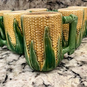 May include: Set of five ceramic mugs shaped like ears of corn. The mugs are yellow and green with a textured surface.