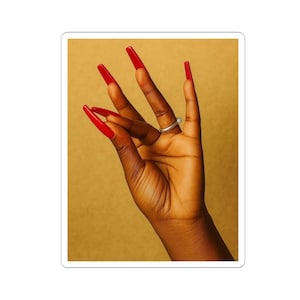 May include: A sticker featuring a hand with long, red fingernails and a silver ring. The hand is positioned against a golden-yellow background, creating a striking contrast between the dark skin tone and the vibrant nail color.