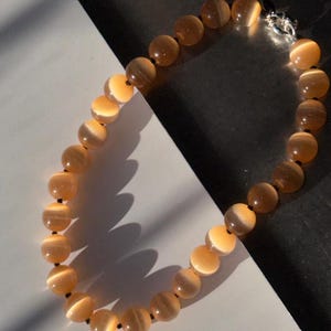 Op de afbeelding: Een ketting met ronde, amberkleurige kralen met een gestreept patroon. De kralen zijn geregen met zwart draad en vastgezet met een zilverkleurige sluiting. De ketting wordt gepresenteerd op een wit en donkergrijs oppervlak.