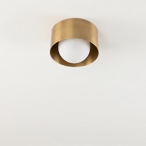 May include: A brass-colored, cylindrical ceiling light fixture with a white, spherical bulb. The light has a simple, modern design, and is mounted on a white ceiling. The fixture is made of metal and has a smooth finish.