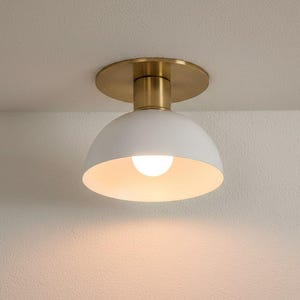 May include: A contemporary ceiling light with a white, domed shade and a brass-coloured base. The light bulb is lit, emitting a warm glow. The fixture is mounted on a white ceiling.