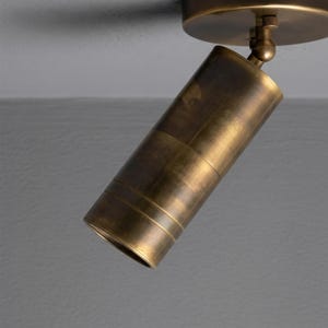 May include: A brass spotlight, cylindrical in shape, attached to a circular base. The spotlight is angled downwards, highlighting its ribbed detailing and adjustable head. Designed for focused illumination.