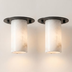 Op de afbeelding: Een paar cilindrische wandlampen met witte, doorschijnende stenen kappen met natuurlijke aders. Elke armatuur is bevestigd aan een ronde, matzwarte metalen basisplaat, wat zorgt voor een warme verlichting.