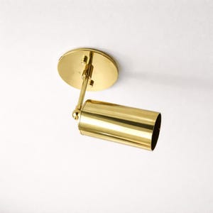 May include: A gold-colored, cylindrical spotlight fixture. The fixture is mounted on a circular base and has an adjustable arm. The spotlight is designed to be mounted on a ceiling and provides focused illumination.