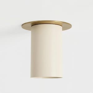 May include: A contemporary ceiling light. It has a cylindrical, cream-coloured shade with a circular, gold-toned trim. The light is designed to be mounted flush against the ceiling, offering a minimalist aesthetic.
