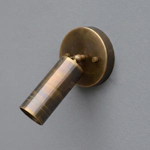 May include: A brass-coloured wall-mounted spotlight. It features a circular base and an angled, cylindrical light head with etched lines. This directional lighting fixture is designed for focused illumination, suitable for various interior spaces.