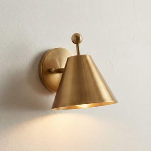 May include: A brass wall sconce featuring a conical lampshade and a circular backplate. The sconce has a small spherical detail at the top and a horizontal arm. The light shines downwards, illuminating the wall below. The sconce is made of brass.