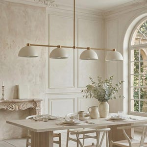 Puede incluir: Un comedor moderno con una mesa rectangular, sillas y una lámpara colgante de cuatro luces. La lámpara tiene un acabado dorado y cuatro pantallas blancas en forma de cúpula. La habitación tiene una paleta de colores neutros con una chimenea y una ventana grande.