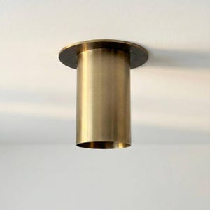 May include: A brass-coloured ceiling light fitting. It features a circular base and a long, cylindrical body. The light is suspended from a white ceiling, casting a soft shadow.