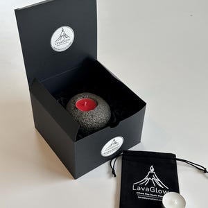 Puede incluir: Una caja de regalo negra contiene un portavelas de piedra gris con una vela roja. Una pequeña bolsa negra con cordón y una vela de té están junto a la caja. La caja y la bolsa tienen el logotipo "LavaGlow".