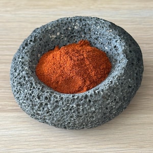 Könnte beinhalten: Eine graue, poröse Steinschale gefüllt mit leuchtend orange-rotem Gewürz. Die Schale hat eine raue, strukturierte Oberfläche und steht auf einer hellen Holzoberfläche. Das Gewürz scheint fein gemahlen zu sein.