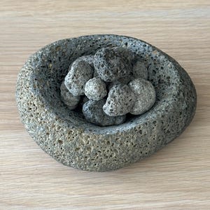 Op de afbeelding: Een grijze, getextureerde stenen kom gevuld met verschillende kleine, poreuze, grijze stenen. De kom heeft een afgeronde vorm met een ruw, ongelijkmatig oppervlak. De stenen zijn in de kom samengebracht.