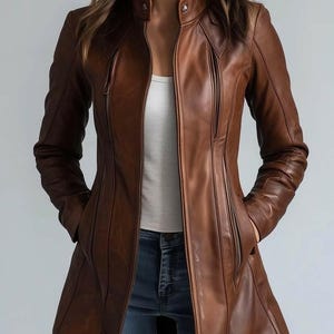May include: A brown leather jacket with a tailored fit, featuring a stand-up collar, multiple zippered pockets, and a front zip closure. The jacket has a slightly flared hem and is worn over a white top and blue jeans.