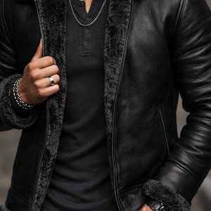 Puede incluir: Chaqueta de cuero negra con cuello y forro de borrego gris oscuro. La chaqueta tiene cierre de cremallera y un diseño clásico. El modelo lleva una camisa gris oscuro y vaqueros negros. Se ve un reloj y una pulsera plateados.
