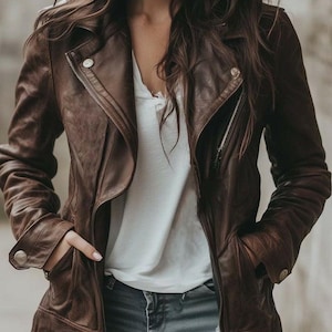 May include: A brown leather jacket with a classic asymmetrical zipper design. The jacket features a notched collar, multiple pockets, and silver-tone hardware. The model is wearing a white shirt and blue jeans.